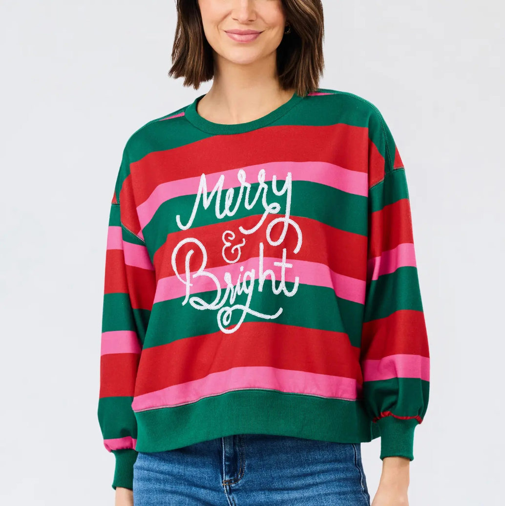 A woman wearing a festive holiday sweatshirt with horizontal red, green, and pink stripes and 'Merry & Bright' written in white script across the front, paired with blue jeans against a light gray studio background.