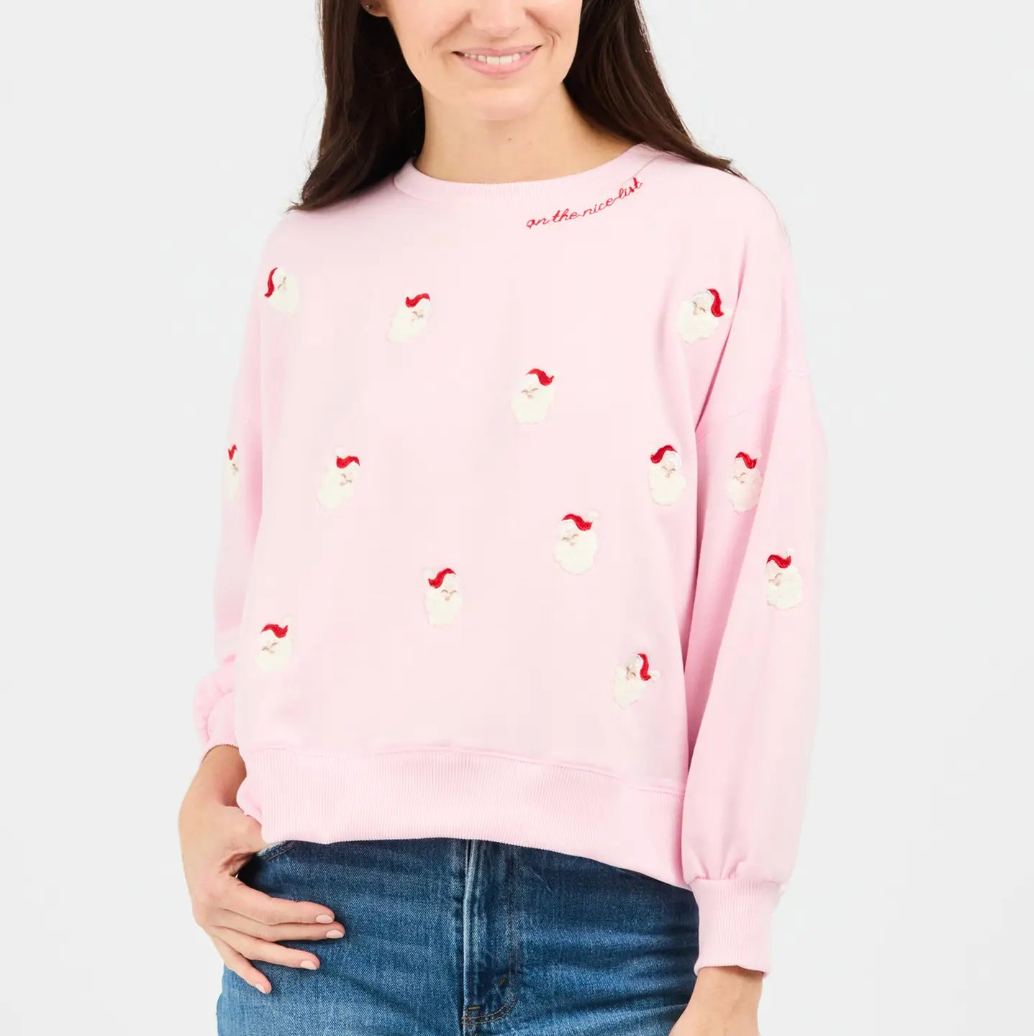 A woman wearing a light pink crewneck sweatshirt decorated with small embroidered snowmen with red hats, paired with blue jeans, against a plain white background.