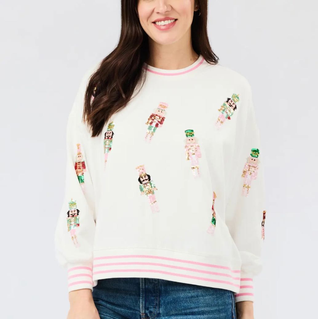 A woman wearing a white crewneck sweatshirt with pink striped ribbed trim and colorful nutcracker graphics, paired with blue jeans against a light gray studio background.