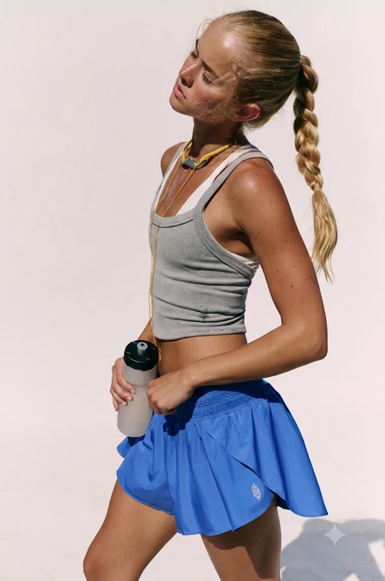 A woman in athletic wear posing outdoors against a light background, wearing a gray ribbed tank top and a bright blue tennis skirt, holding a water bottle.