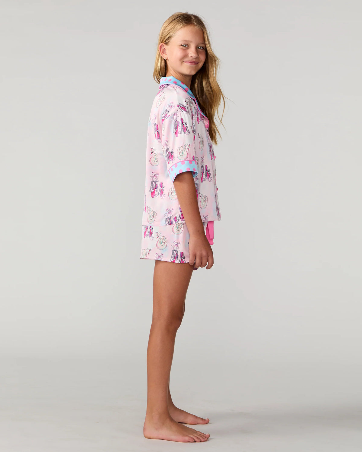 A young girl stands barefoot in profile wearing a light pink pajama set with a colorful cartoon pattern and light blue trim on a plain light gray studio background.