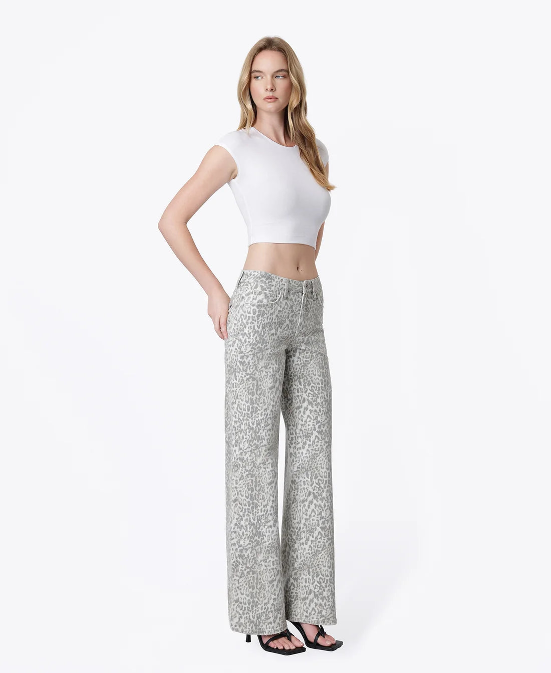 A woman modeling a white cropped short-sleeve top and high-waisted, wide-leg trousers with a silver and gray leopard print, paired with black sandals, against a plain white studio background.