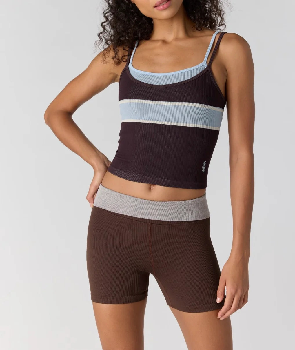 A woman modeling a dark brown ribbed two-piece activewear set featuring a cropped tank top with light blue and white horizontal stripes, and matching high-waisted shorts with a light gray waistband.