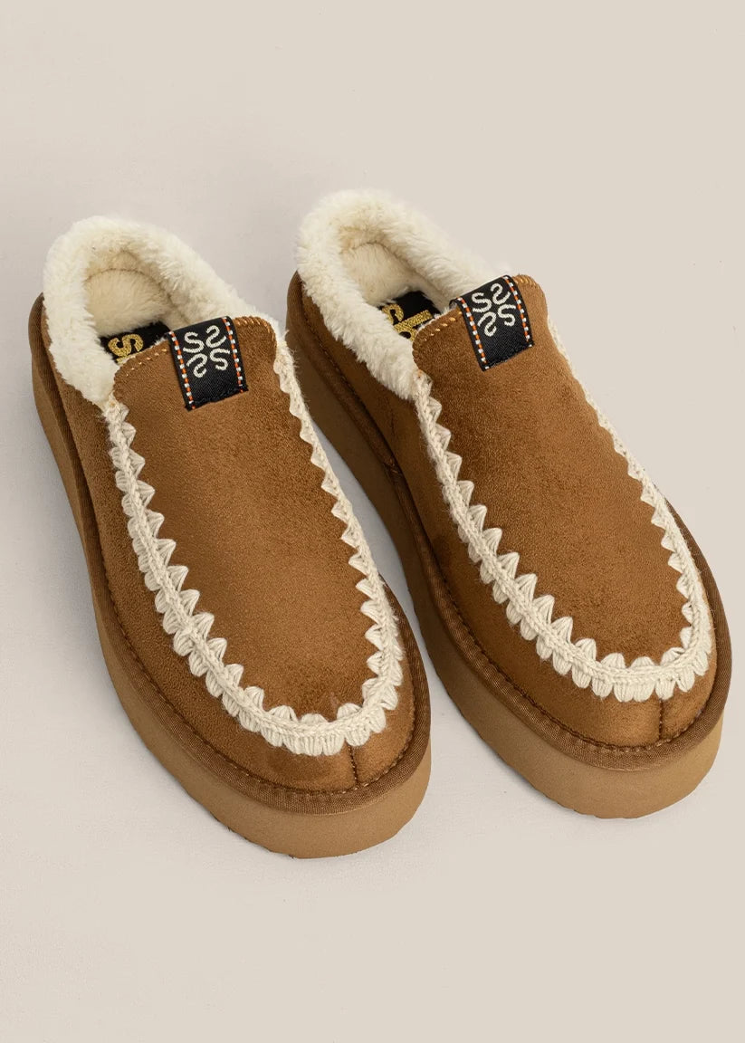 A pair of brown suede slip-on slippers with white faux-fur lining and a decorative white braided trim, shown on a light beige background.
