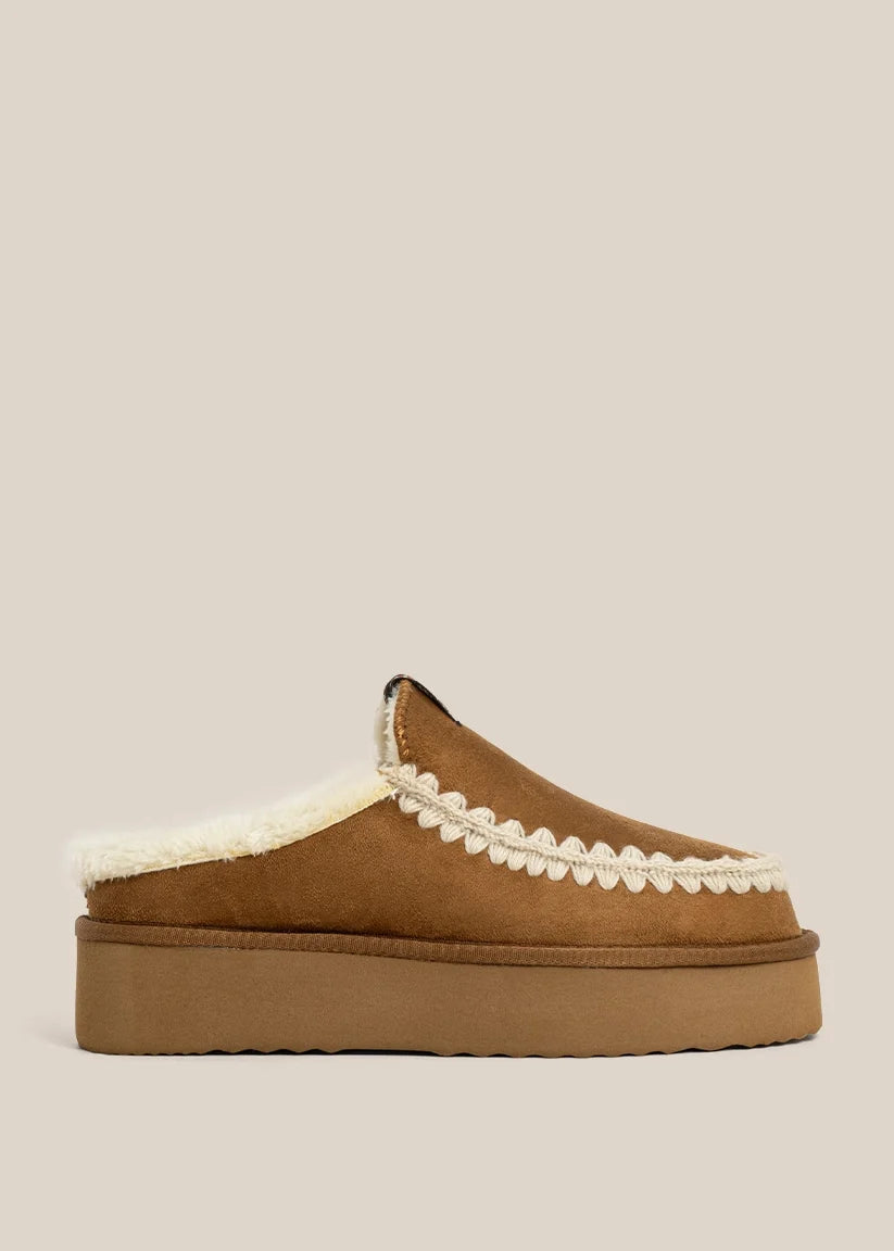 A single brown suede slip-on mule slipper with a thick platform sole, cream shearling lining, and a decorative cream scalloped trim, shown against a solid light beige background.