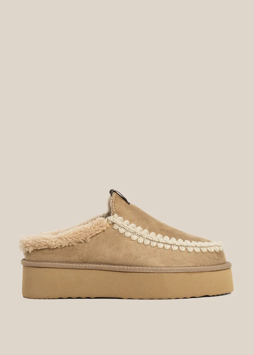 A single platform slip-on mule shoe in tan suede with a cream sherpa lining and a decorative cream scalloped trim, shown against a solid light beige background.