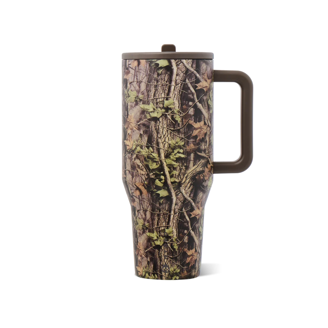A travel tumbler with a brown handle and lid, featuring a realistic woodland camouflage pattern of tree bark, branches, and green leaves, shown on a white background.