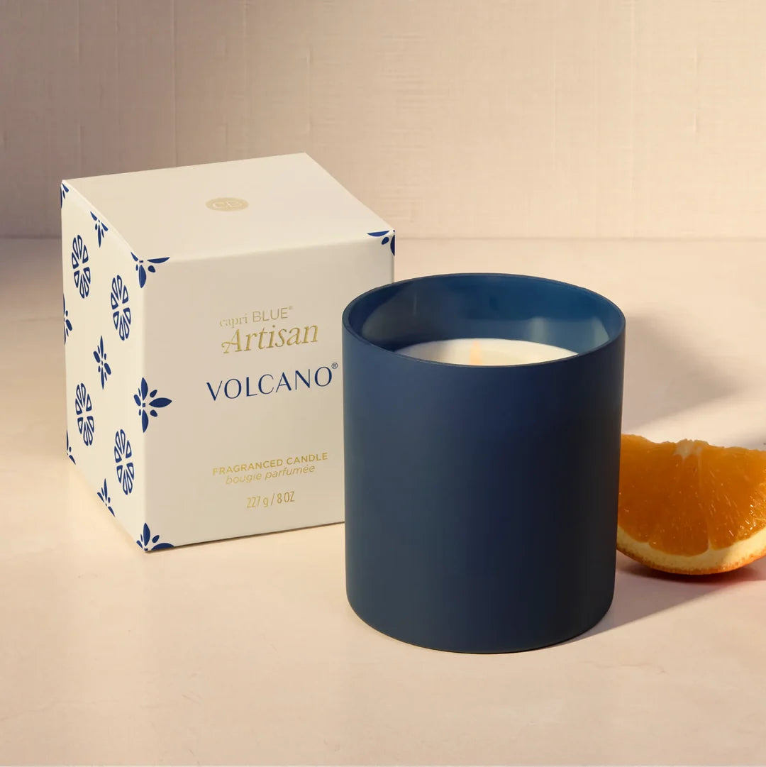 A matte navy blue glass candle jar with white wax sits next to its white and blue patterned retail box, with a slice of orange on a warm beige surface.