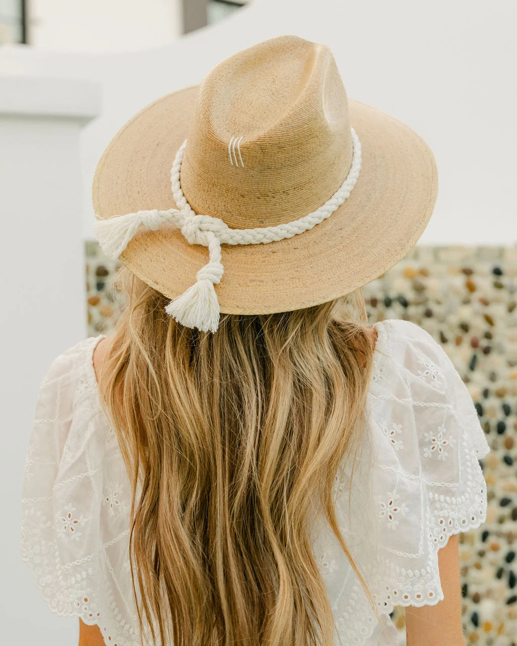 A woman viewed from behind wearing a light beige straw fedora hat with a white braided ribbon and tassel, paired with a white eyelet blouse.