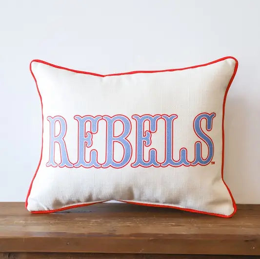 A square decorative throw pillow with a cream linen-like fabric, red piping, and the word 'REBELS' embroidered in blue with a red outline across the front, resting on a wooden surface against a white wall.