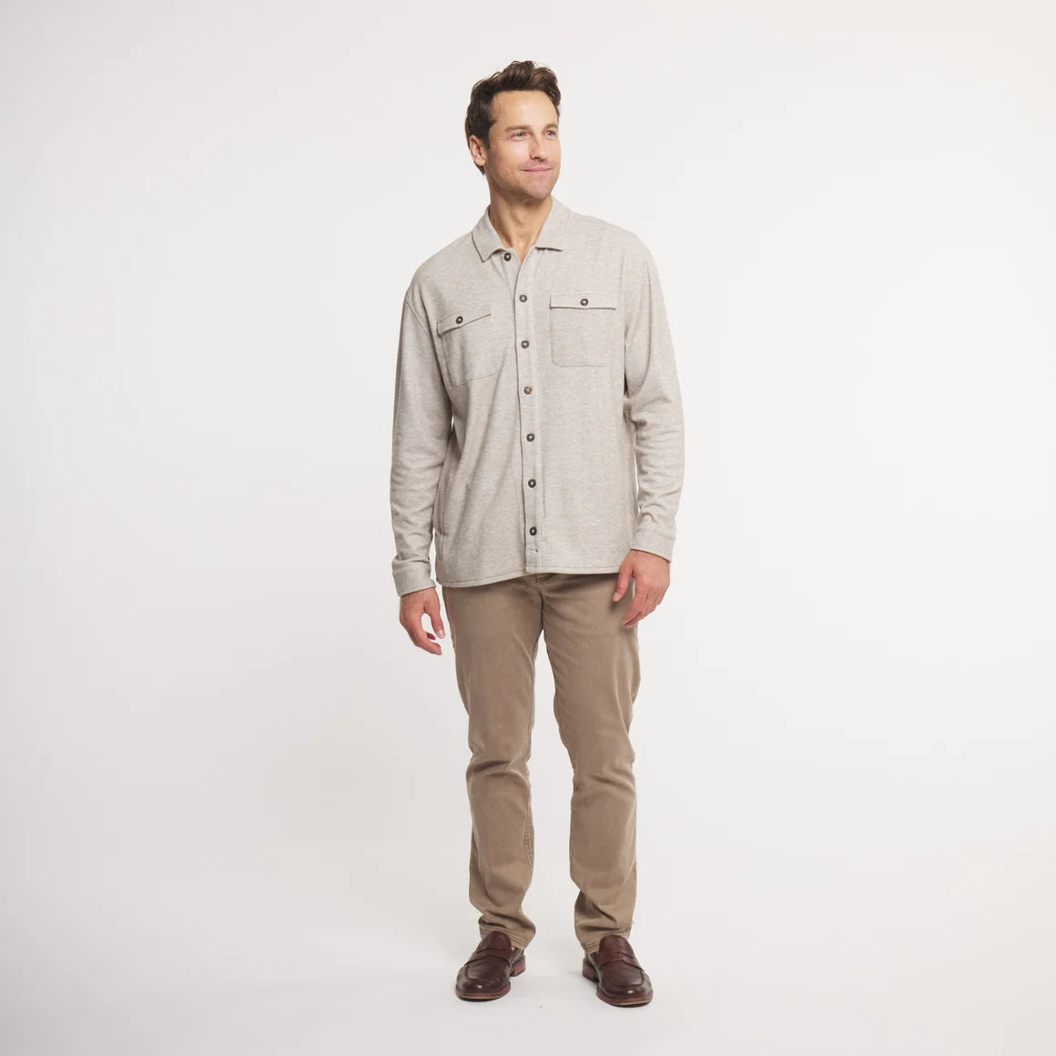 A man wearing a light beige long-sleeve button-up shirt with two chest pockets and tan pants, standing against a plain white studio background.