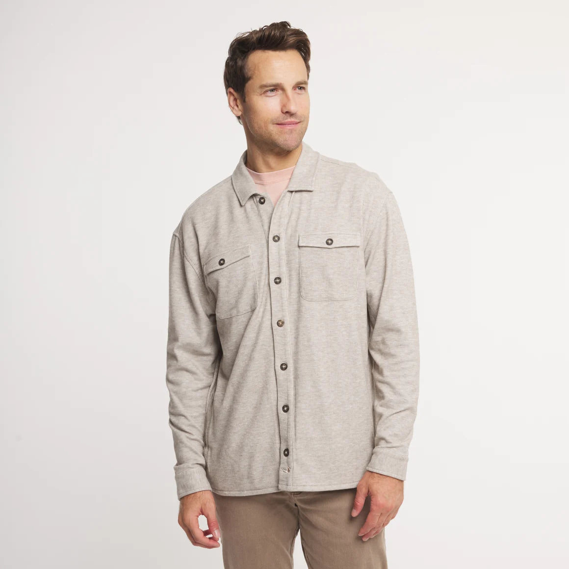 A man wearing a light beige heathered long-sleeve button-up shirt with two chest pockets, paired with tan pants, standing against a plain white studio background.