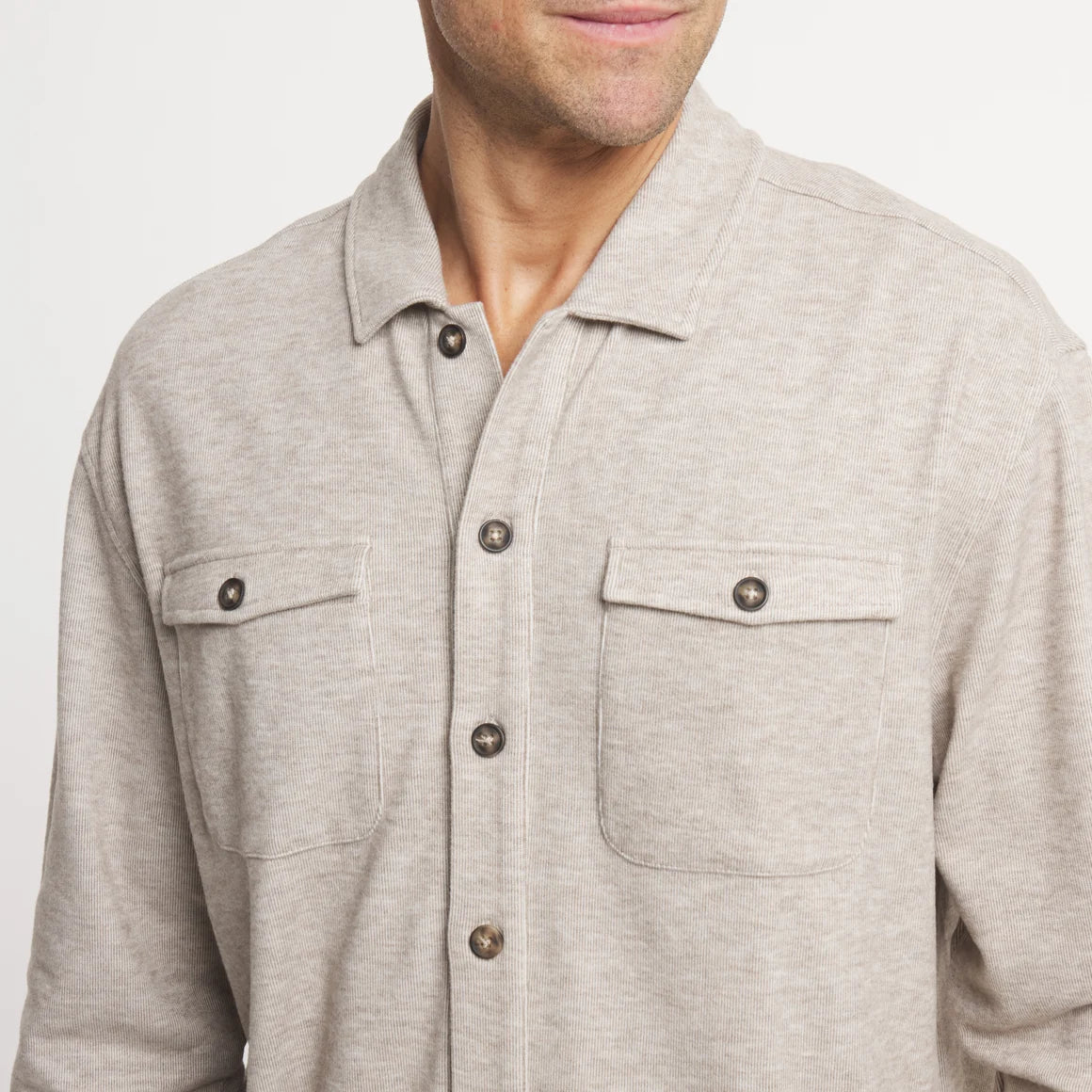 A close-up of a man wearing a light heather gray long-sleeve button-up shirt with two chest pockets and dark buttons, against a plain light background.