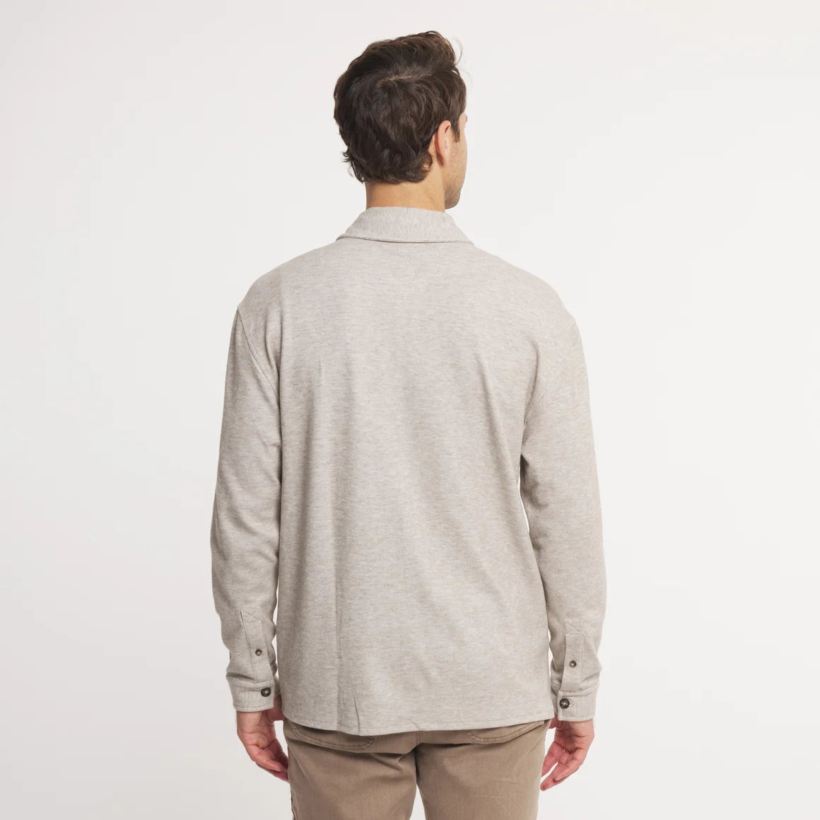 A man viewed from the back wearing a light heather gray long-sleeve knit polo shirt and tan trousers, set against a plain white studio background.