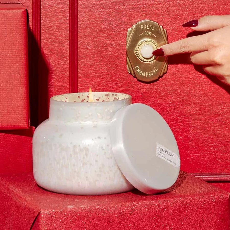 A white speckled glass candle jar with its lid leaning beside it on a red surface, with a hand pressing a champagne bottle stopper-style button against a red textured background.