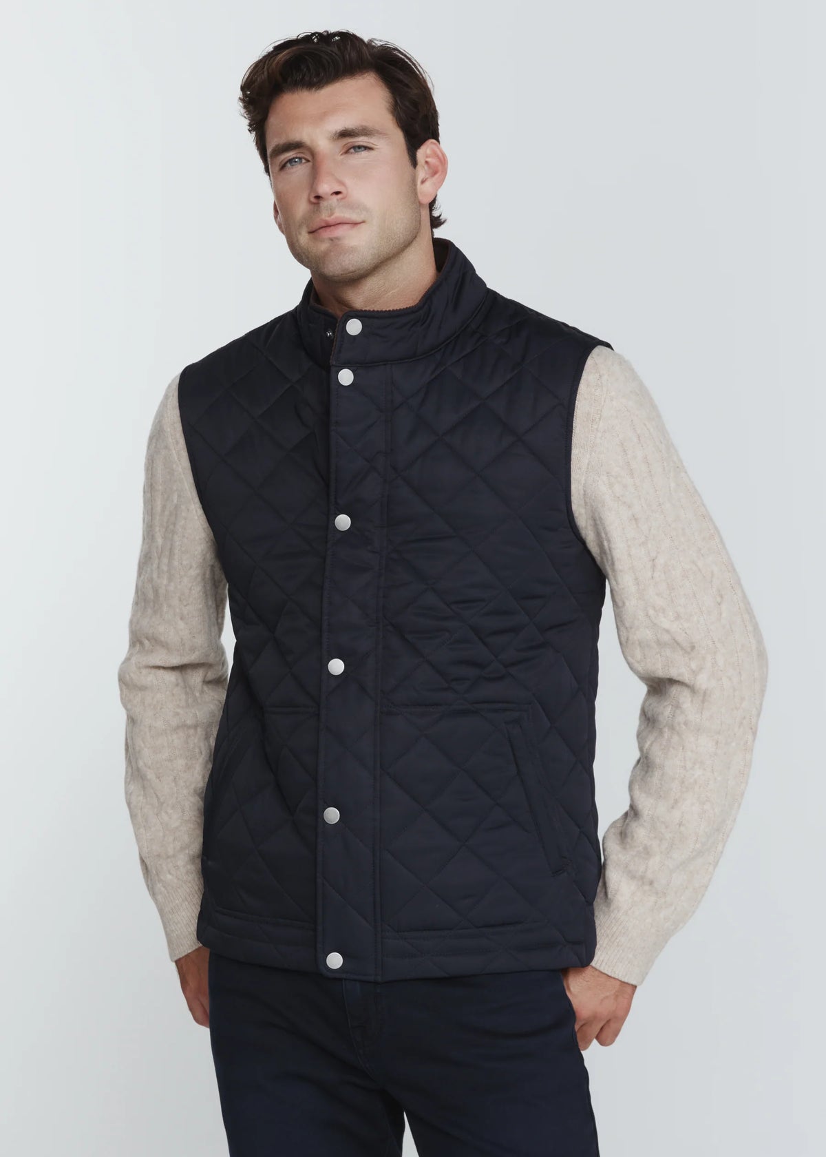 A man wearing a navy blue diamond-quilted button-up vest over a beige knit sweater and dark trousers, photographed against a light gray studio background.