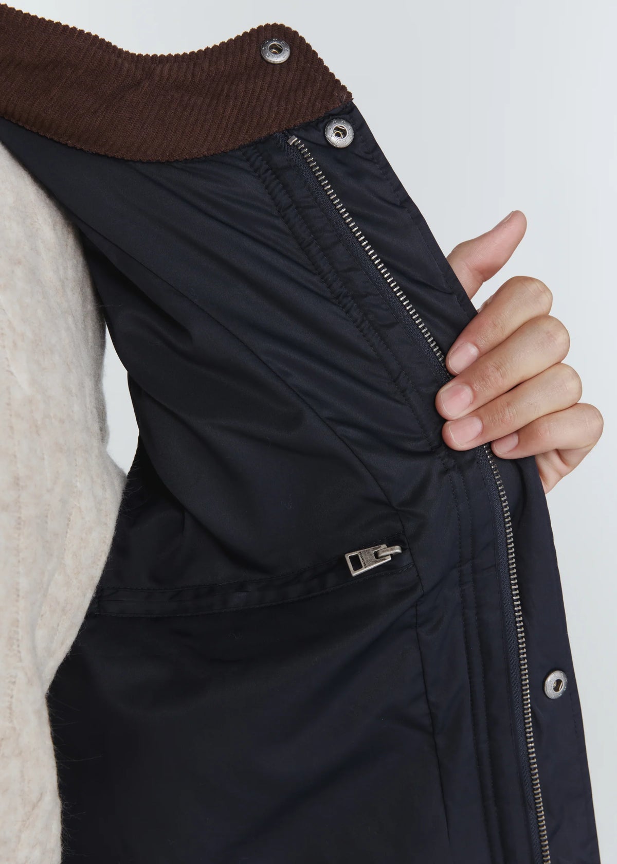 Close-up of a black quilted jacket with a brown corduroy collar, silver snap buttons, and a hand holding open a front zipper.