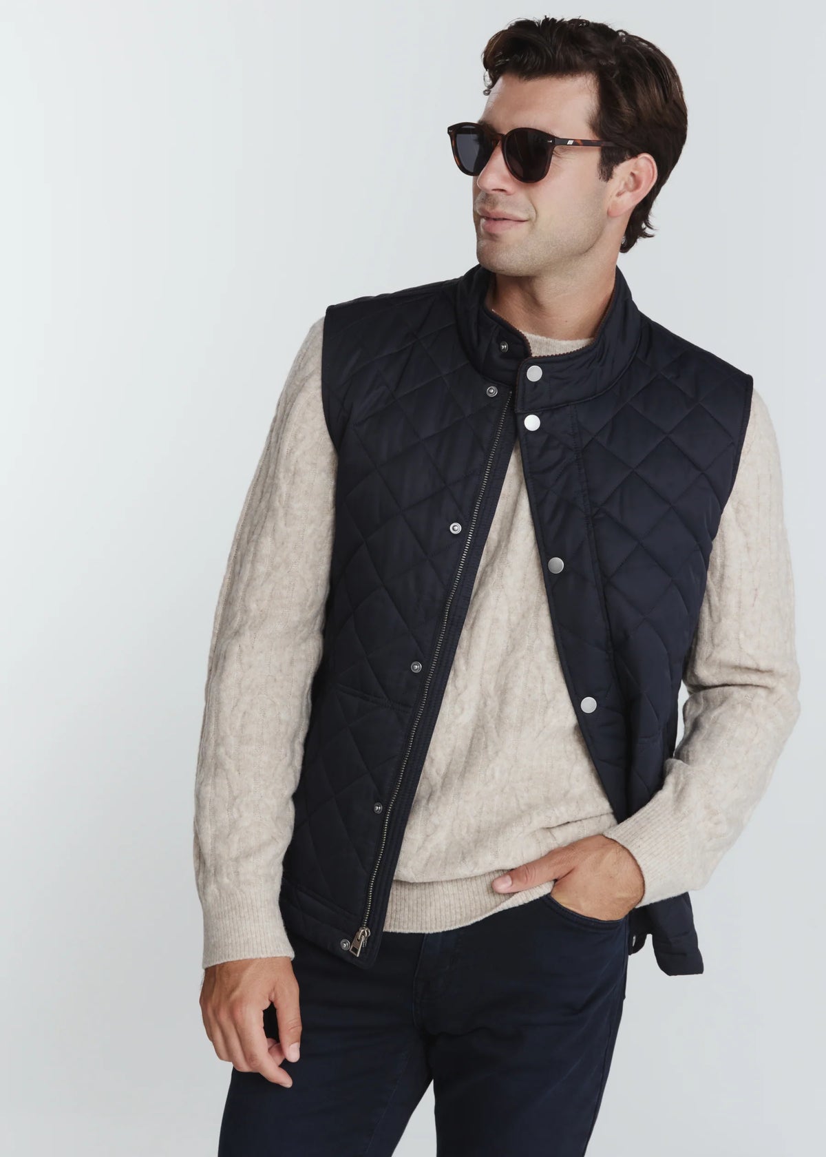 A man wearing a dark navy quilted zip-front vest over a light beige knit sweater and dark pants, photographed against a clean light gray studio background.