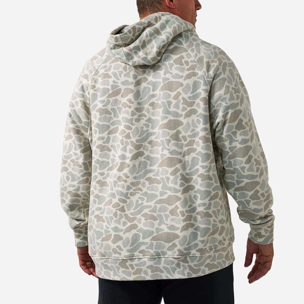 A man viewed from the back wearing a light sage green and beige camouflage hoodie, paired with black pants, against a plain light gray background.