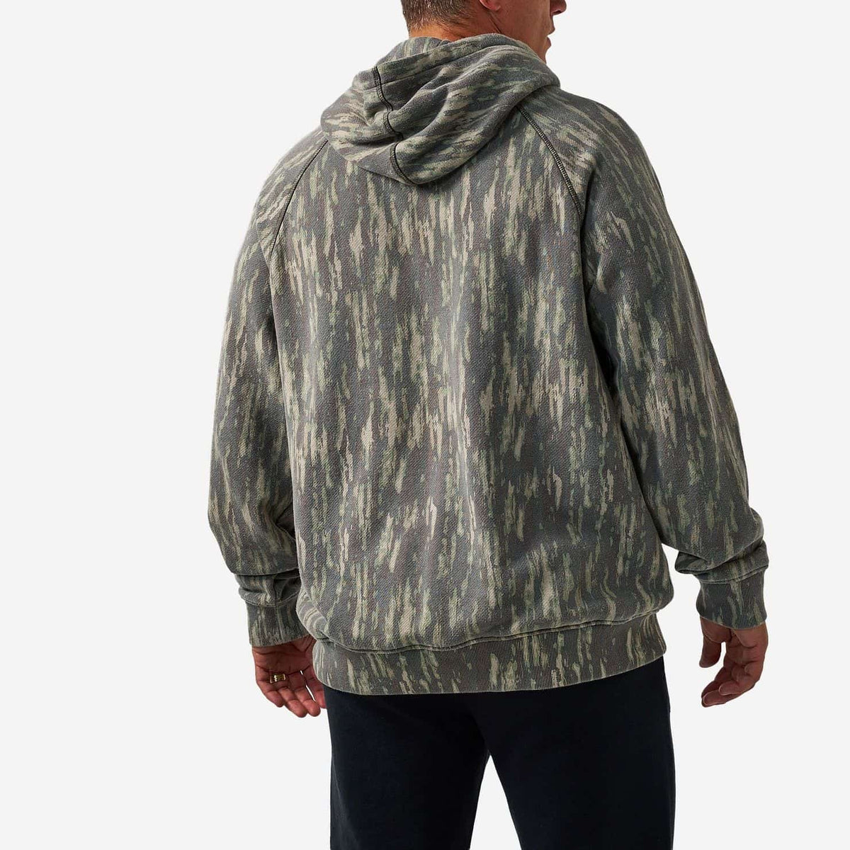 A man viewed from the back wearing a faded olive green and gray hooded sweatshirt with a distressed, mineral-wash pattern, paired with black pants, against a light gray studio background.