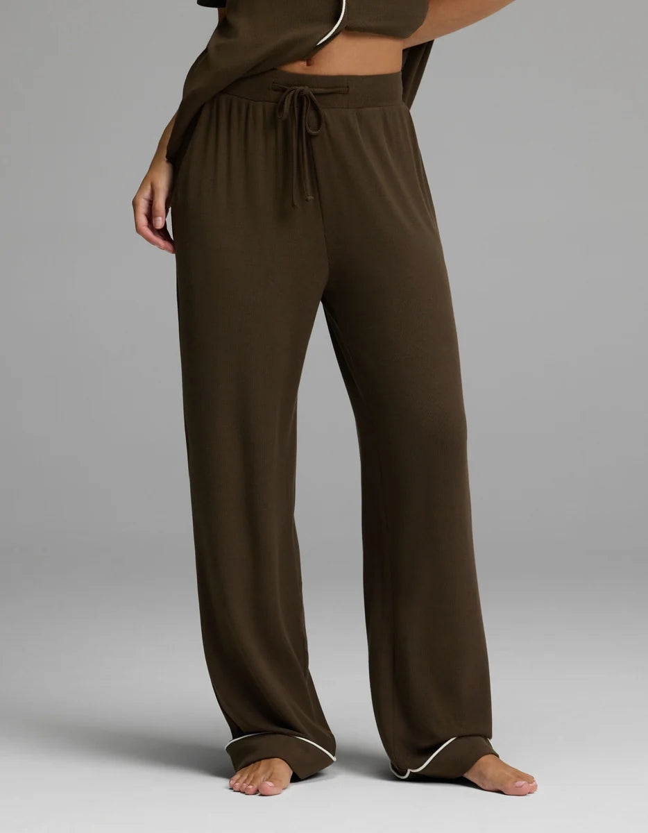 A person wearing loose, wide-leg drawstring pajama pants in dark brown, shown from the waist down against a light gray studio background.