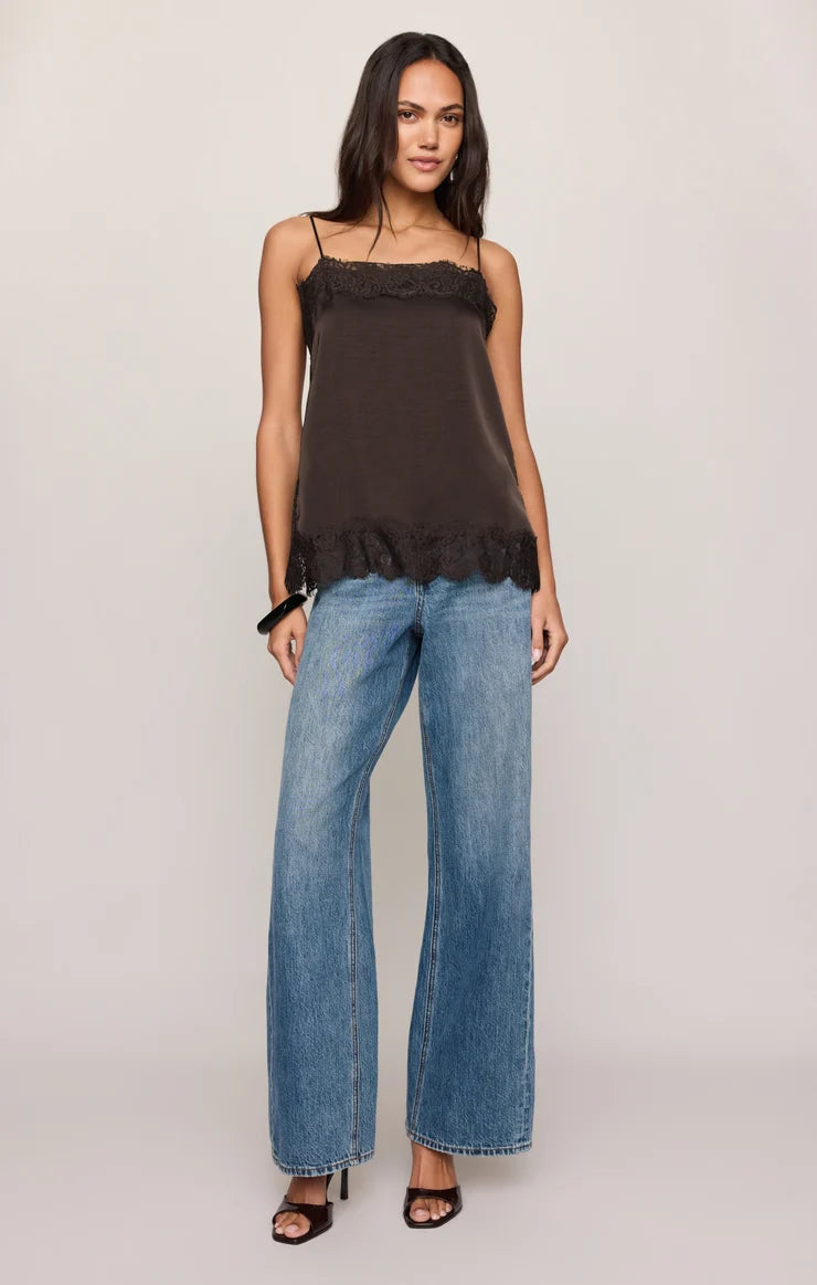 A woman modeling a dark brown camisole top with delicate lace trim at the neckline and hem, paired with wide-leg blue jeans and black heeled sandals, against a plain light gray studio background.