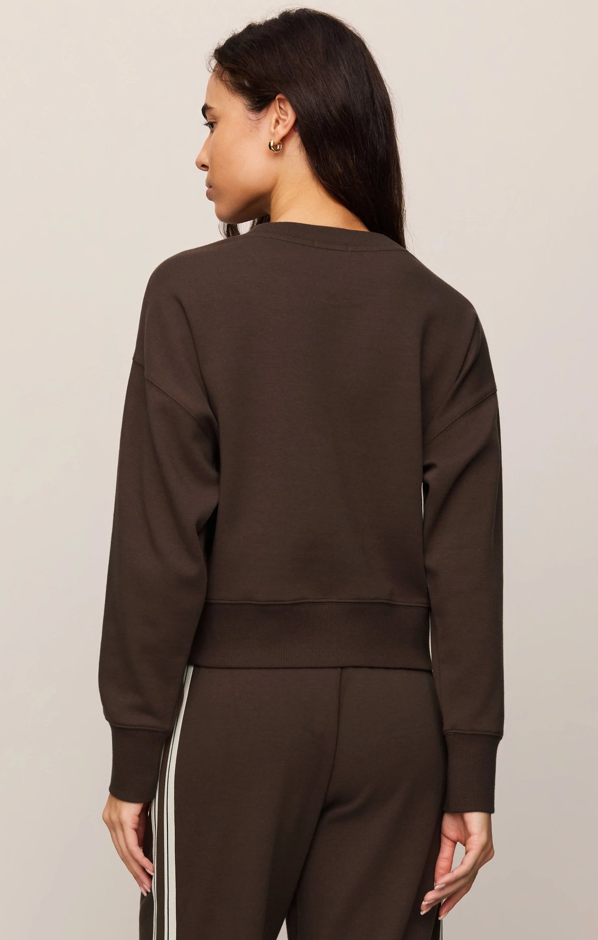 A woman shown from the back wearing a dark brown long-sleeve cropped sweatshirt and matching dark brown pants with white side stripes, against a plain light beige studio background.