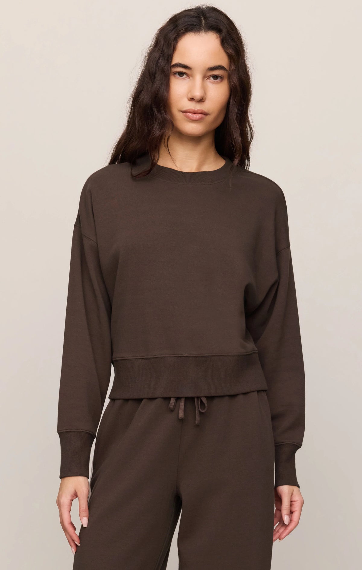 A woman modeling a dark brown cropped long-sleeve sweatshirt and matching drawstring sweatpants against a plain light gray studio background.