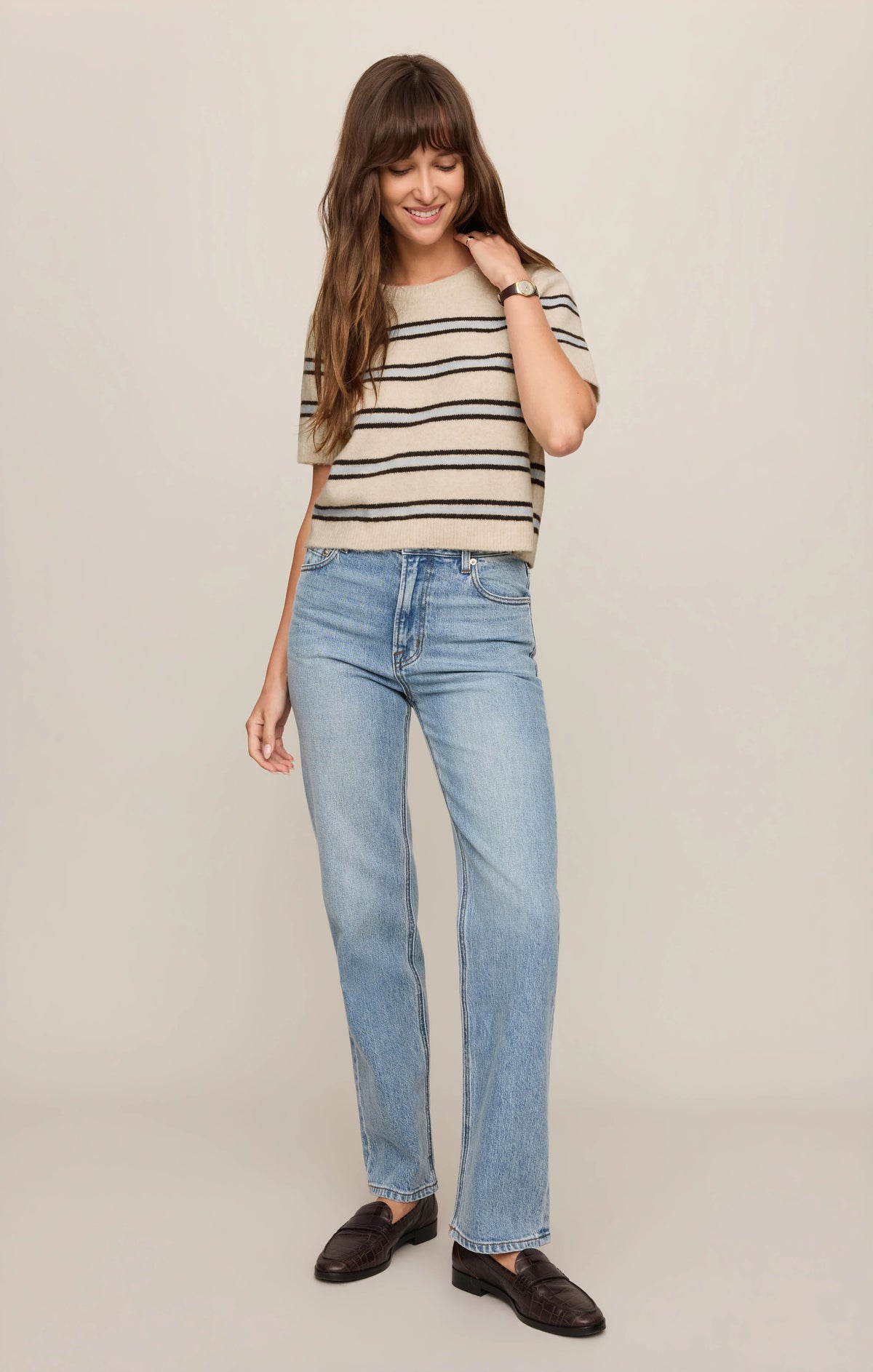A woman modeling a beige short-sleeve knit top with thin black horizontal stripes and light-wash blue jeans, paired with dark brown loafers, against a plain light beige studio background.