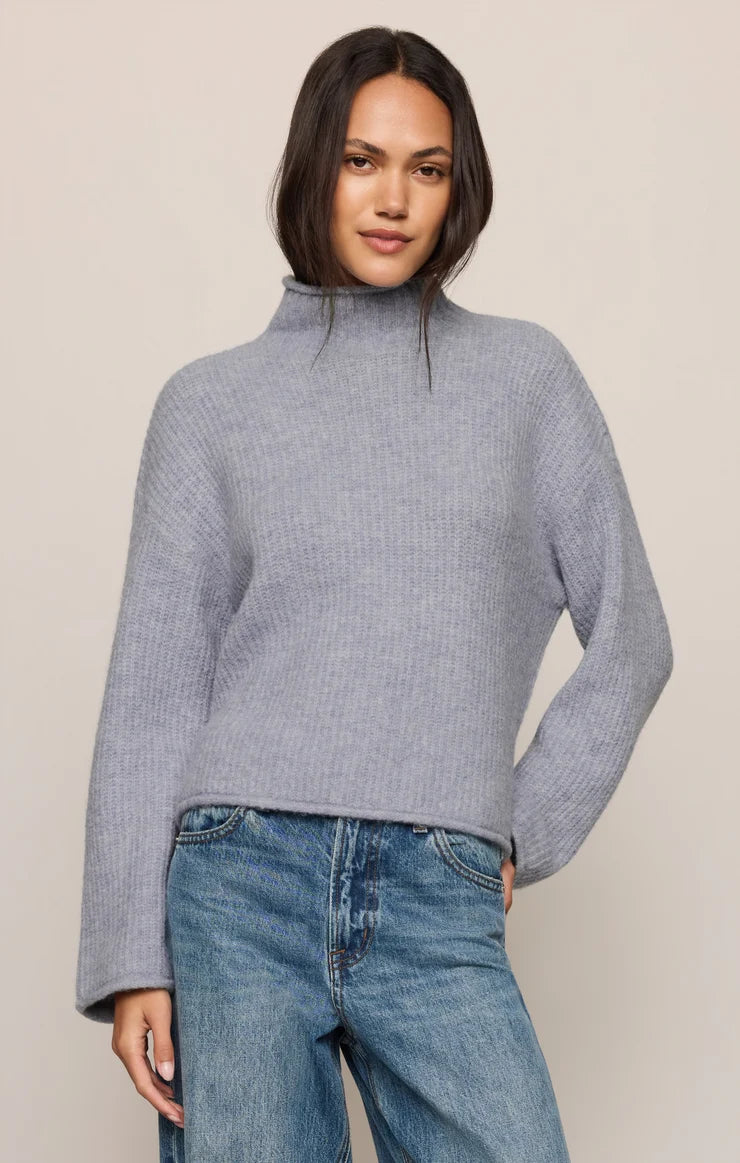A woman wearing a light gray ribbed knit mock neck sweater and blue jeans, standing against a plain beige studio background.