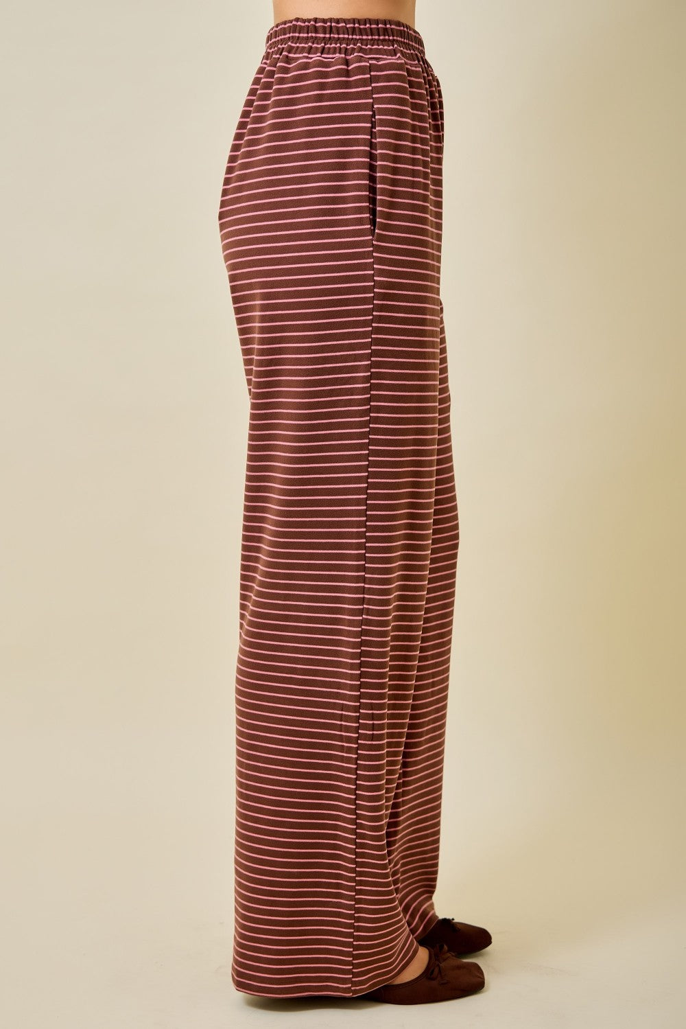 A person shown from the waist down wearing loose, wide-leg pants with thin horizontal stripes in rust red and white, paired with dark brown slip-on shoes, against a plain beige studio background.