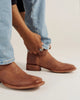 A person wearing light brown leather cowboy boots with light blue jeans, adjusting the ankle strap of the right boot. The boots feature a square toe and a low block heel.
