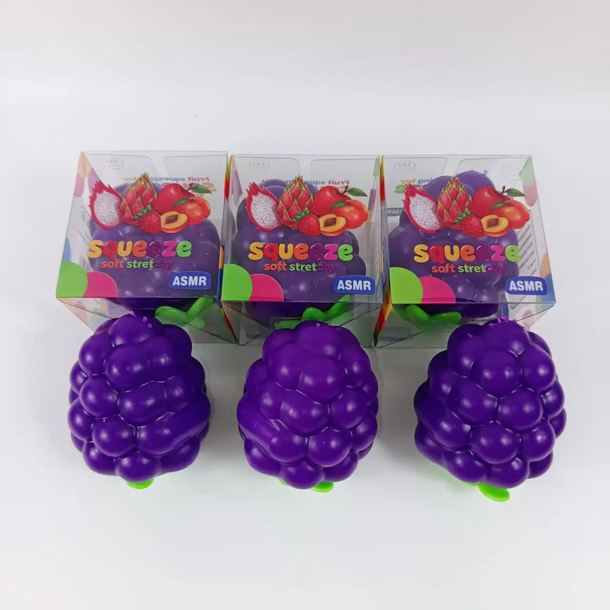 Three purple grape-shaped squeeze toys in front of their clear plastic packaging boxes, which feature colorful fruit illustrations and the brand name ASMR.