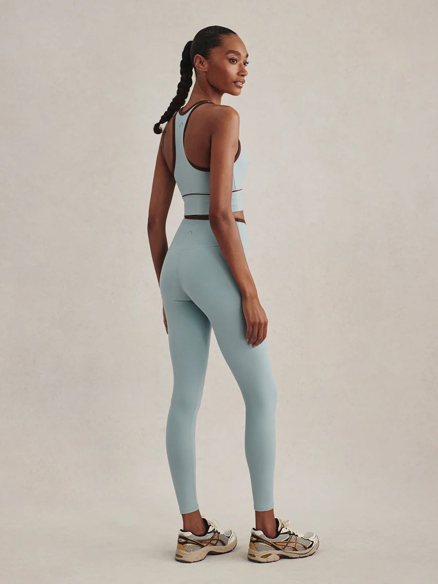 A woman modeling a matching light blue activewear set consisting of a sports bra and high-waisted leggings, paired with chunky sneakers, against a neutral beige studio background.