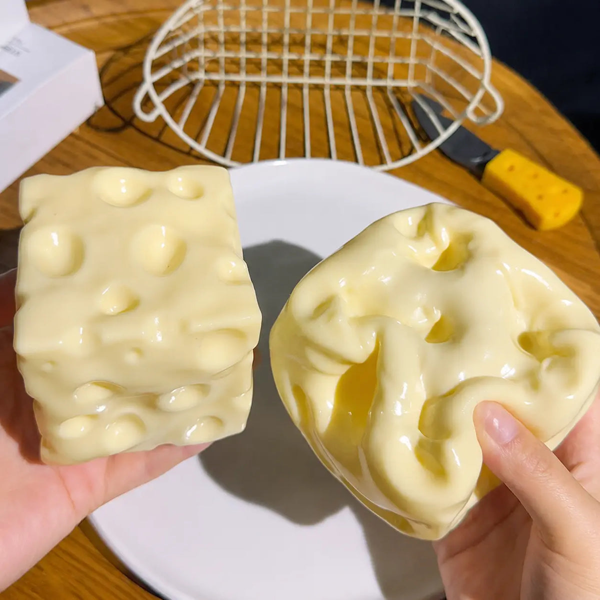 Two hands holding cheese-shaped novelty items with a glossy pale yellow surface and raised holes, shown over a white plate on a wooden table with a wire rack and knife in the background.