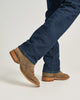 A close-up of a person's legs wearing brown suede cowboy boots and dark blue denim jeans, captured from the side against a clean, light gray studio background.