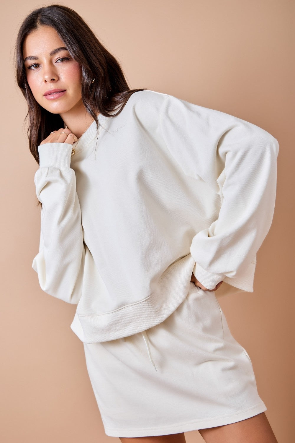 A woman modeling an oversized off-white sweatshirt and matching sweat shorts against a solid beige studio background.