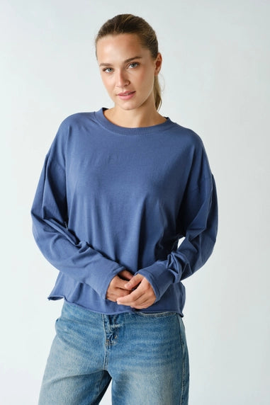 A woman wearing a slate blue long-sleeve crewneck top and light wash blue jeans, standing against a light gray studio background.