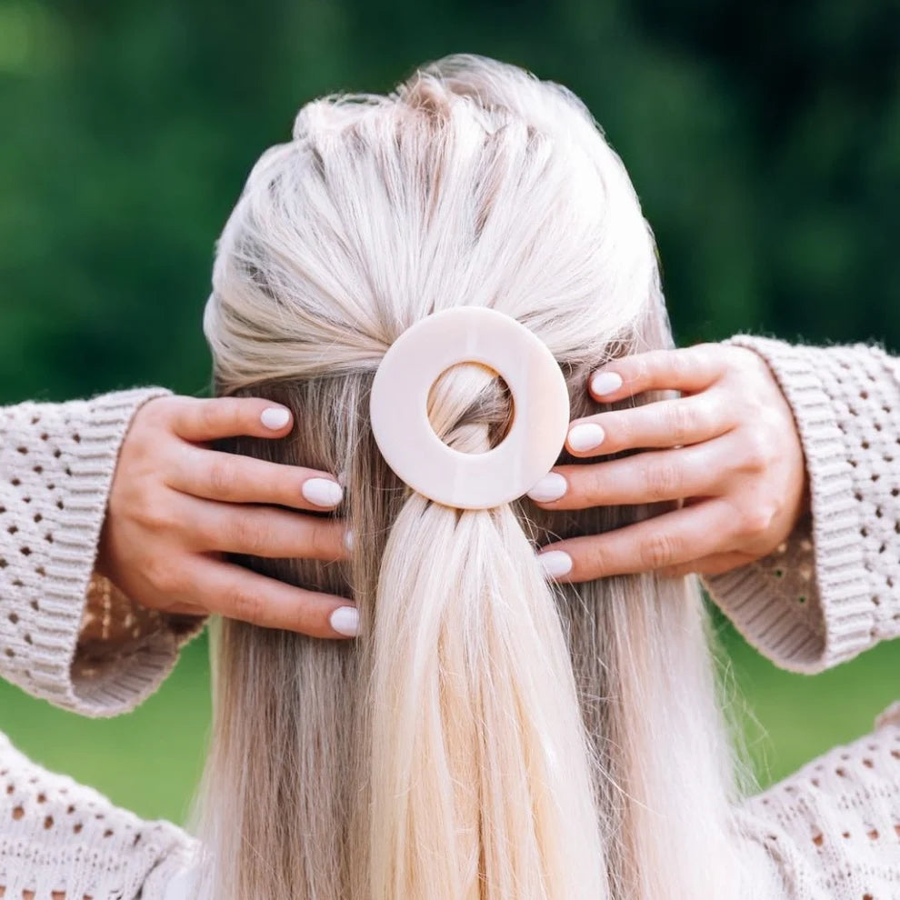 A person viewed from behind wearing a large, circular, pale pink hair tie in their light blonde hair, with their hands adjusting it. The background is a soft, blurred green outdoor setting.