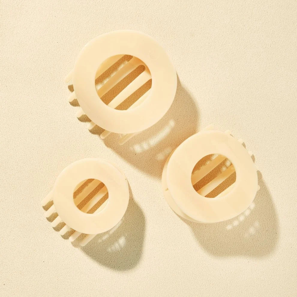 Three cream-colored plastic hair claw clips arranged on a light beige textured background with soft shadows.