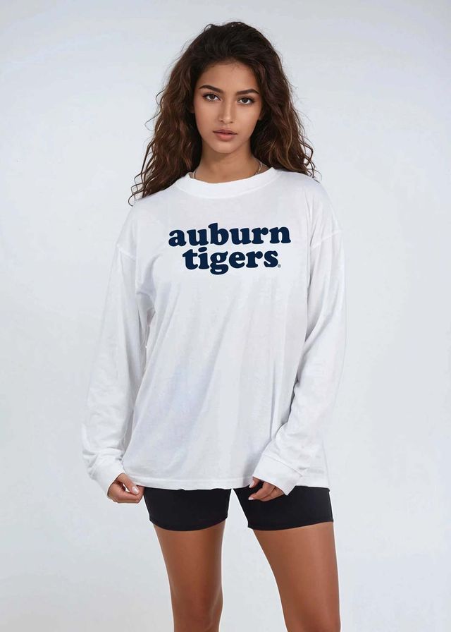 A woman modeling a white long-sleeve t-shirt with 'auburn tigers' printed in navy blue across the chest, paired with black athletic shorts, against a light gray studio background.