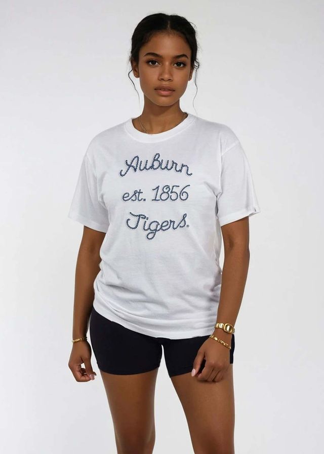 A woman modeling a white short-sleeve t-shirt with 'Auburn est. 1856 Tigers' printed in navy blue script on the front, paired with black athletic shorts, against a plain light gray background.