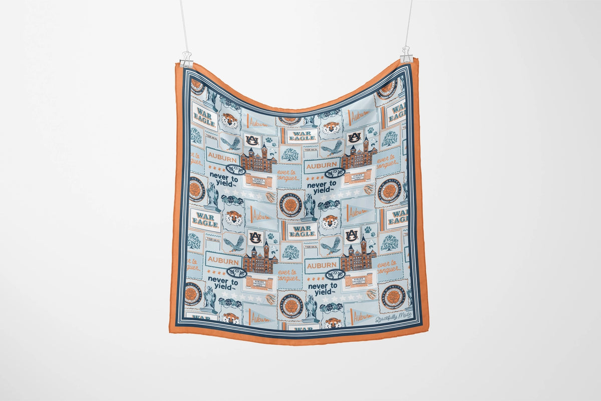 A square fabric scarf with an orange border and a light blue central print featuring vintage-style travel posters, birds, and text, hanging against a plain white wall.