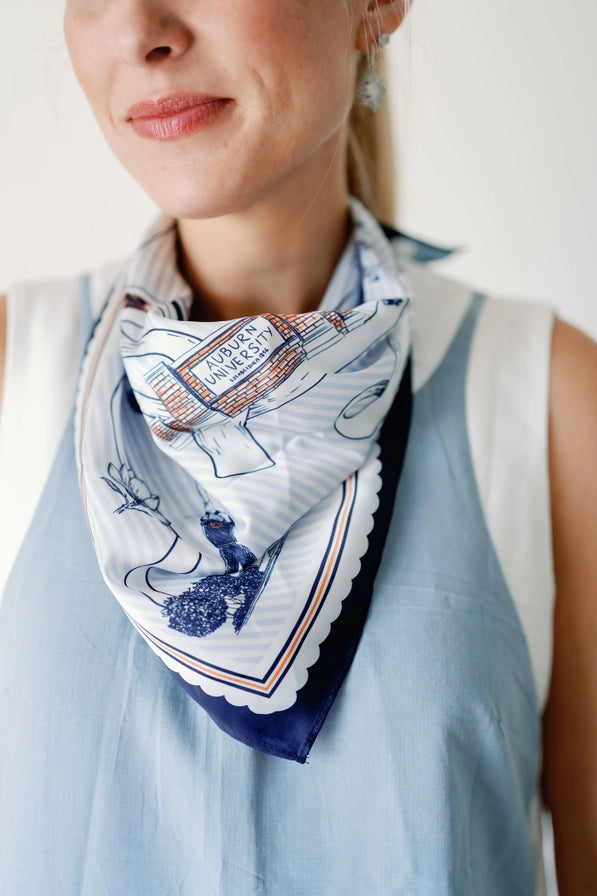 A close-up of a woman wearing a light blue sleeveless top and a patterned white scarf with navy blue illustrations and a scalloped border.