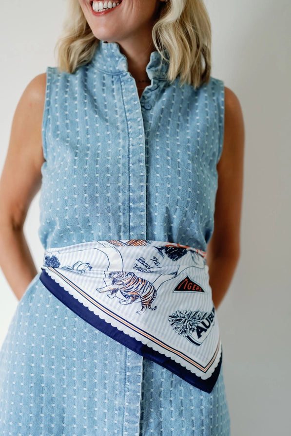 A woman wearing a sleeveless light blue patterned shirt dress with a white scarf featuring a tiger print and navy blue trim tied around her waist.