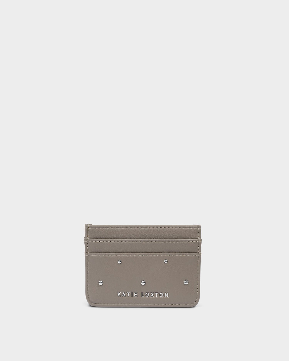 A small taupe leather card holder wallet by Katie Loxton, featuring silver-tone metal studs and the brand name printed in white at the bottom center, set against a solid light gray background.