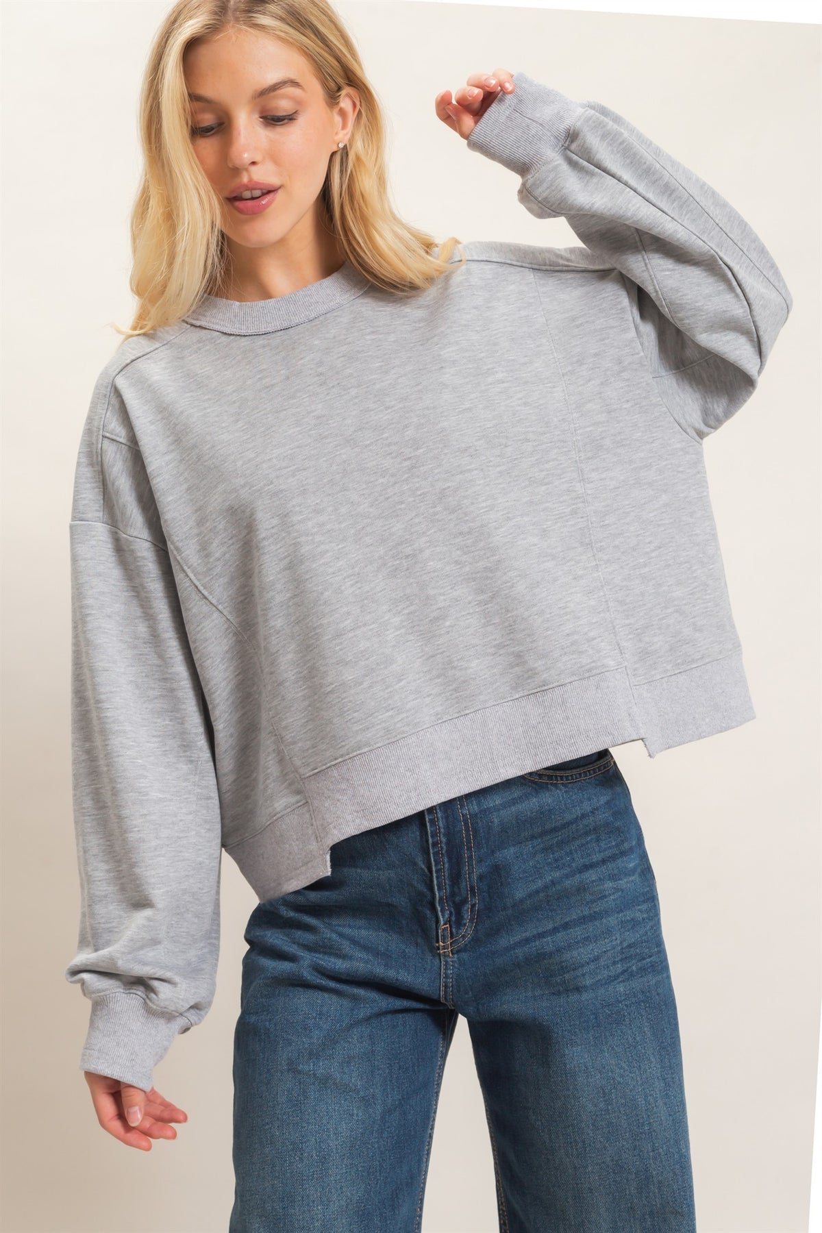 A woman wearing a light gray cropped crewneck sweatshirt and blue jeans, posing against a plain off-white studio background.