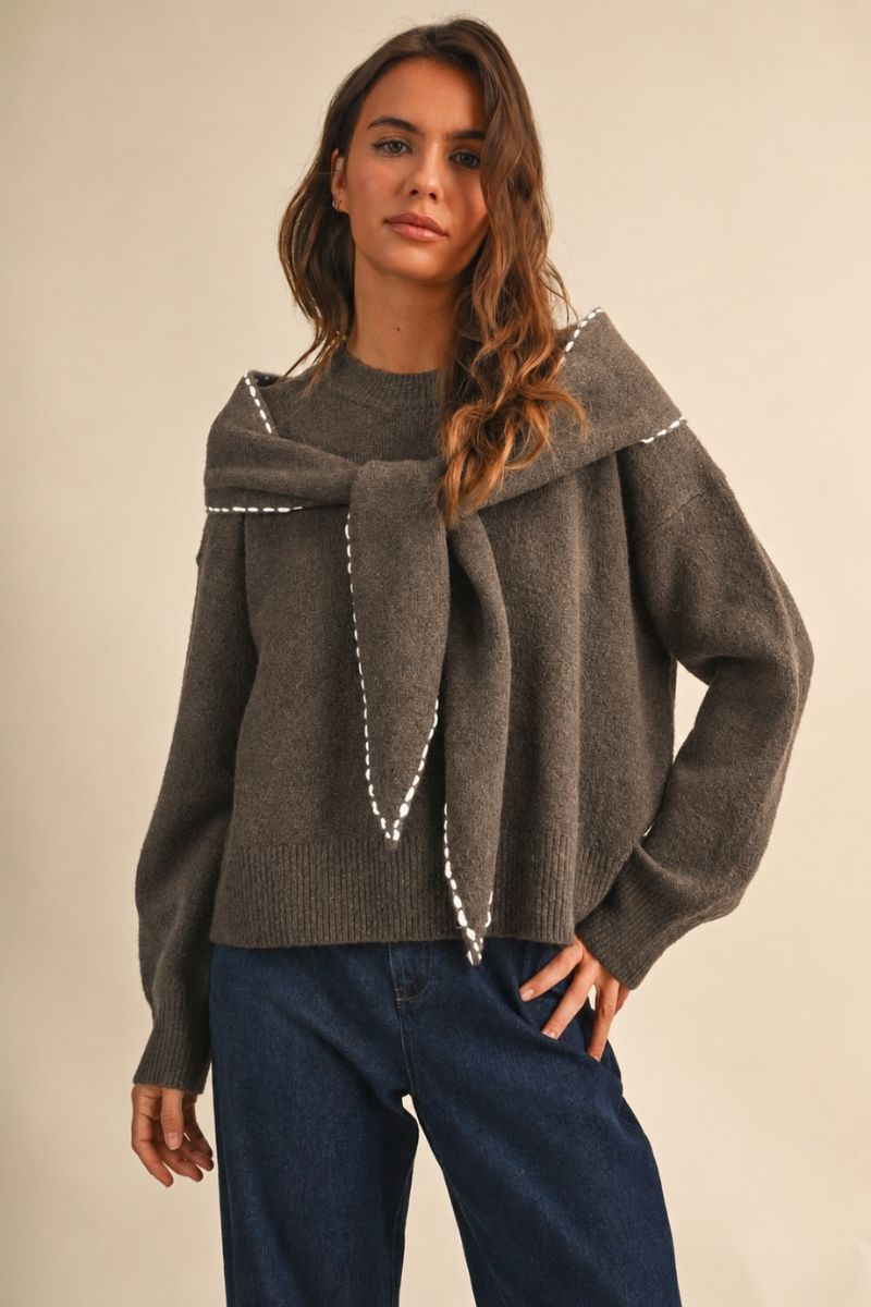 A woman wearing a dark brown knit sweater with a wide collar and a large bow detail outlined with white beadwork, paired with dark blue jeans, against a plain beige background.