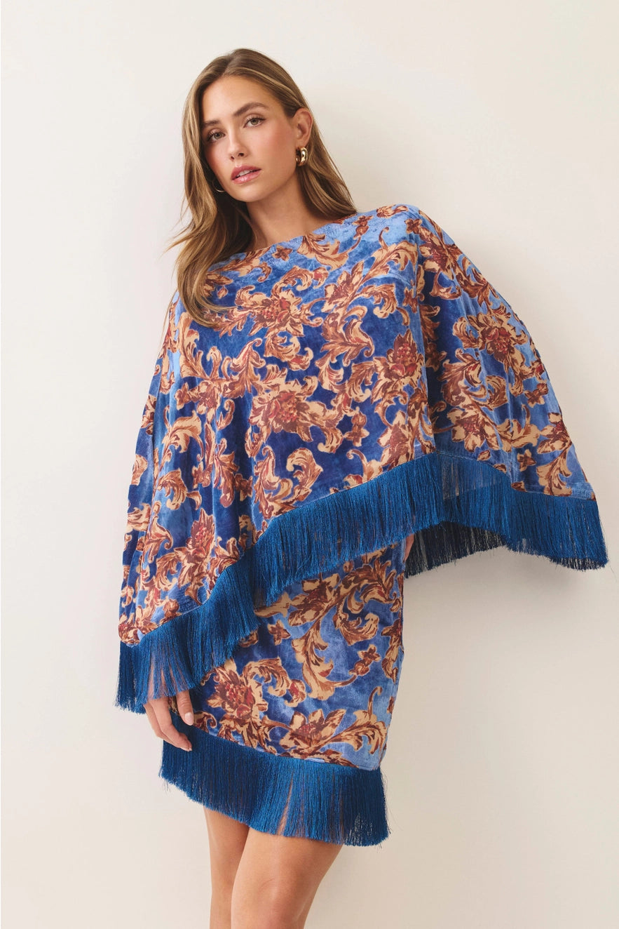 A woman models a blue and brown baroque-patterned poncho with fringed edges against a plain off-white background.
