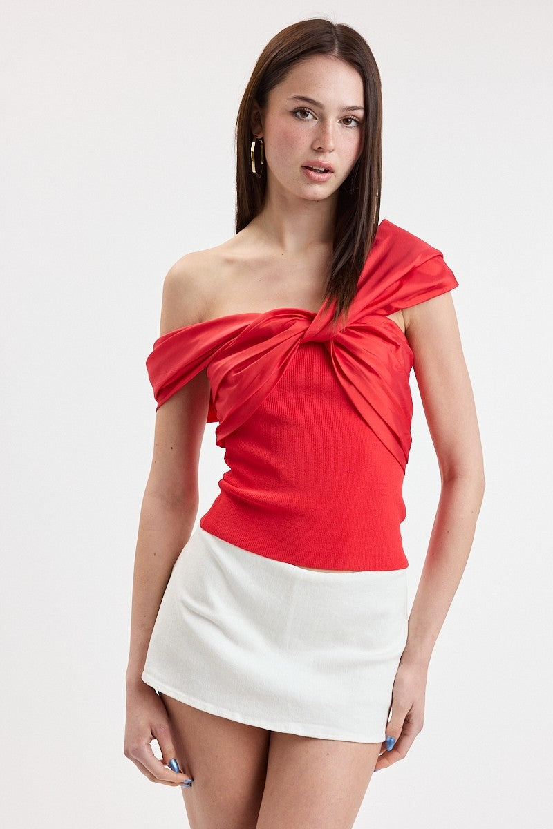 A woman modeling a bright red off-the-shoulder knit top with a large twisted knot detail at the bust, paired with a white mini skirt, against a plain light gray studio background.